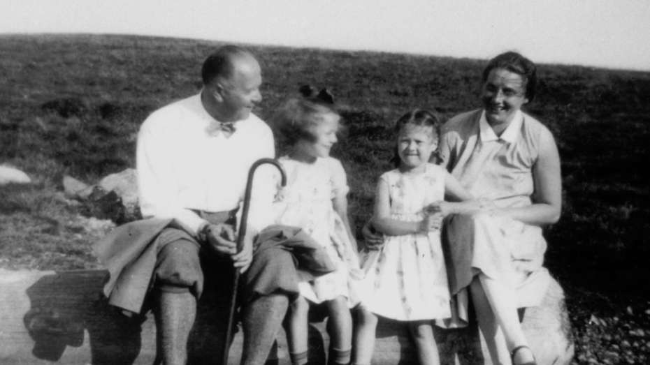 Familie Heimerich, um 1927. Links Hermann, die zwei Kinder in der Mitte und rechts Anneliese. Sie sitzen auf einem Baumstamm. Dahinter ist Wiese zu erkenne. Alle wirken fröhlich. Der Mann hat eine Jacke über den Beinen und einen Wanderstock in der Hand.