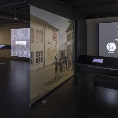 A visitor sits in the left half of the room, and on the wall is the writing "Where you burn books, you also burn people in the end". On screens next to it, one can read the word "Coordination" (Gleichschaltung). In the right half of the room, children can be seen running across a schoolyard. A swastika flag waves on the wall, the screen next to it informs about "Education and Youth under National Socialism".
