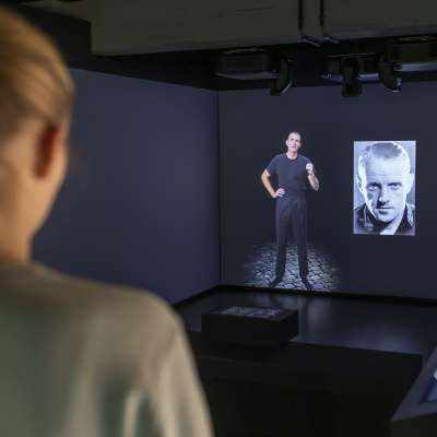 Looking over the shoulder of a blonde young woman at a screen with an actor. In this room, perpetrator and victim biographies are presented in videos.