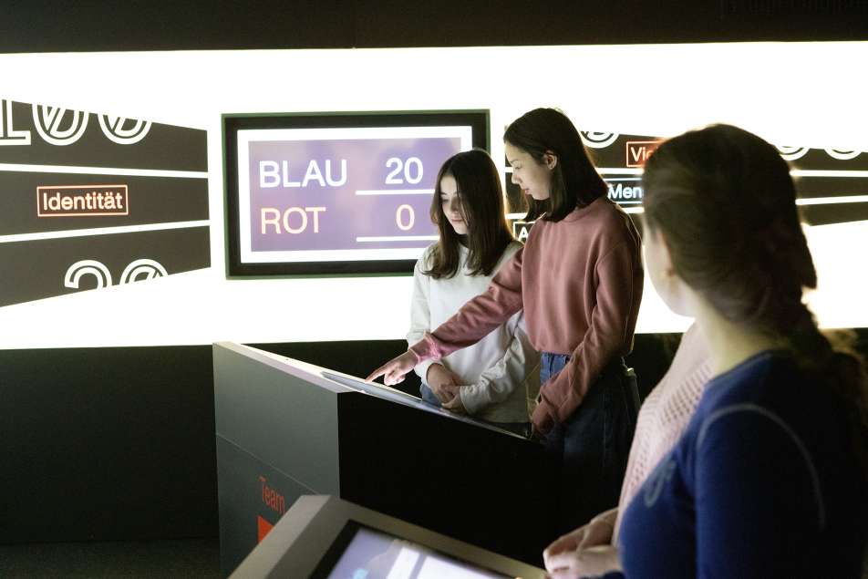 Fotografie von drei jungen Menschen in einem Ausstellungsraum vor Bildschirmen mit Touchscreens