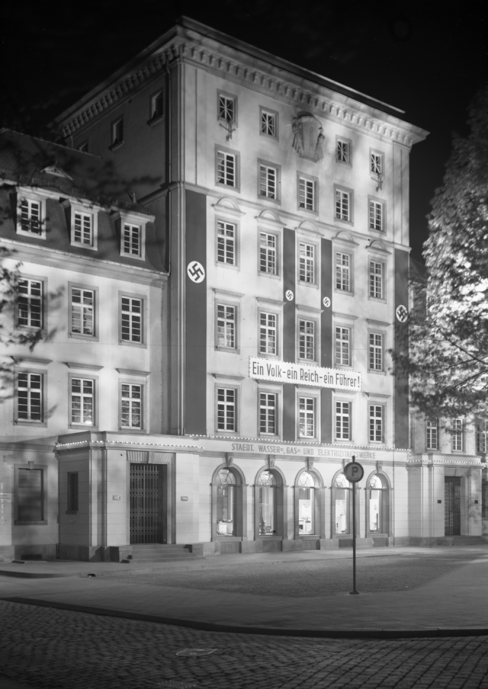 Fotografie in schwarz-weiß zeigt eine beleuchtete Hausfront mit nationalsozialistischen Fahnen bei Nacht
