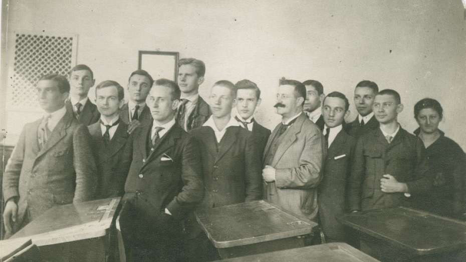 Die Unterprima U I b des Karl-Friedrich-Gymnasiums 1917; Hans Jacobsohn ist der vierte von rechts. 13 Jugendliche mit Lehrkraft stehen ernst blickend in einem Klassenzimmer. Im Vordergrund sind die Schultische zu sehen, dahinter stehen die Schüler teils seitlich gedreht, teils frontal zur Kamera gewandt.
