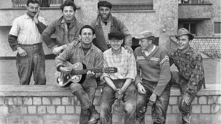 Ein schwarz-weißes Bild. Vier Gastarbeiter sitzen auf einer Mauer, der ganz links hat eine Gitarre in der Hand, die Runde sieht fröhlich aus, alle schauen in die Kamera. Dahinter stehen weitere drei Personen, die ebenfalls lachen.