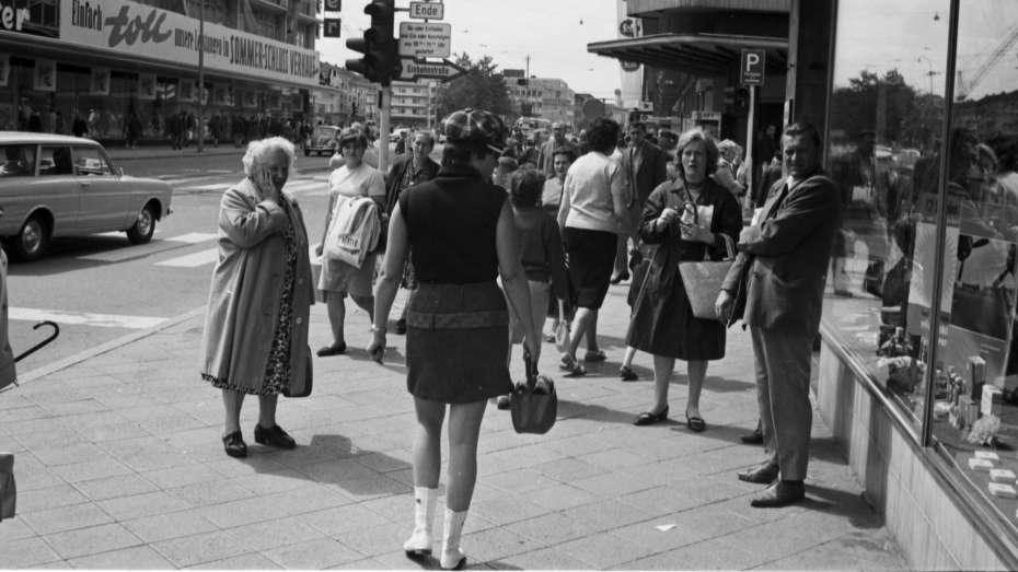 Eine Straßenszene: Eine junge Frau im Minirock läuft in den 1960ern in der Innenstadt. Eine ältere Dame hält sich die Hand vor Erschrockenheit vor den Mund, jüngere Beobachterinnen scheinen interessiert zu schauen.
