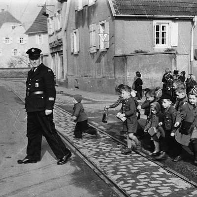 1950 - Mit der Verkehrserziehung kann gar nicht früh genug begonnen werden. Ein Kindergarten wird von einem Polizeibeamten über die Straße geleitet. Seckenheim, Hauptstraße. rechts kath. Kirche St. Aegidius 