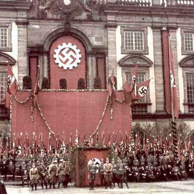 Coloriertes Bild einer nationalsozialistischen Propagandaveranstaltung im Ehrenhof des Schlosses. Das Schloss ist pompös mit Ehrenkränzen, Hakenkreuzen und Hakenkreuzfahnen geschmückt. Davor stehen Nazis in Festuniform.