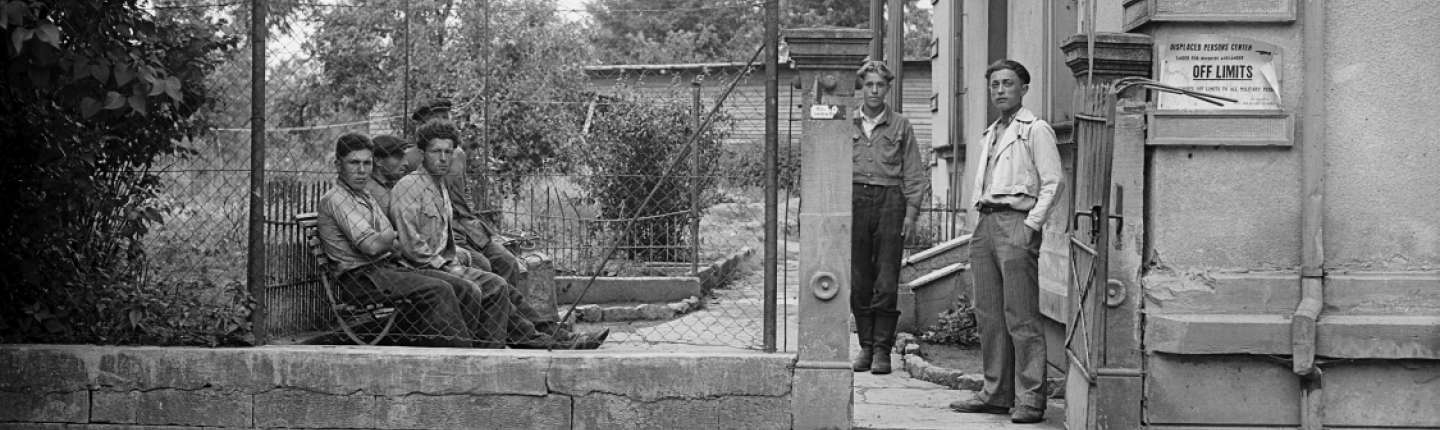 Fünf Menschen in einem abgezäunten Gelände einer Sammelunterkunft im Jahre 1946.