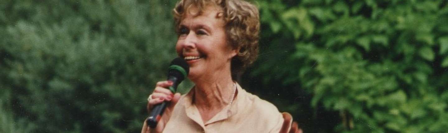 Farbiges Foto von einer älteren Frau namens Elsbeth Janda mit Mikrophon auf einer Bühne mit Bäumen im Hintergrund