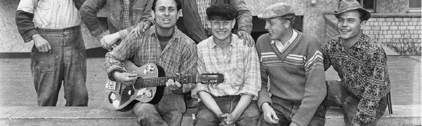 Ein schwarz-weißes Bild. Vier Gastarbeiter sitzen auf einer Mauer, der ganz links hat eine Gitarre in der Hand, die Runde sieht fröhlich aus, alle schauen in die Kamera. Dahinter stehen weitere drei Personen, die ebenfalls lachen.