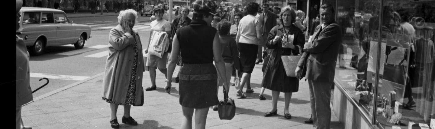 Eine Straßenszene: Eine junge Frau im Minirock läuft in den 1960ern in der Innenstadt. Eine ältere Dame hält sich die Hand vor Erschrockenheit vor den Mund, jüngere Beobachterinnen scheinen interessiert zu schauen.