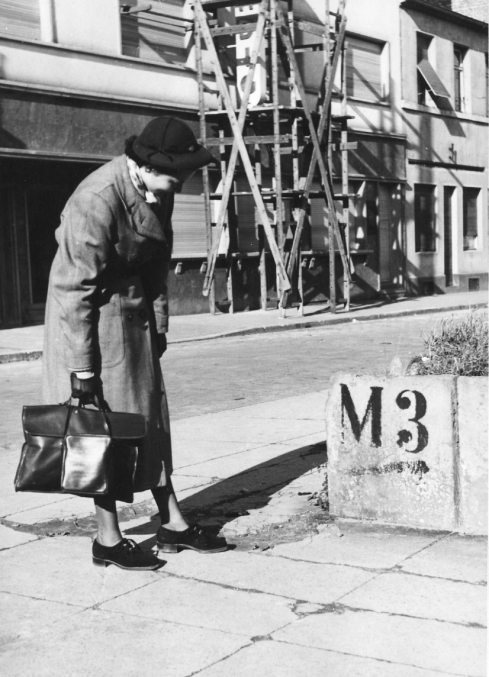 Fotografie in schwarz-weiß zeigt eine Frau mit Hut, Mantel und Tasche auf einer Straße in den Mannheimer Quadraten. Die Person steht leicht gebückt auf de linken Seite und schaut sich einen Mauerrest mit der Bezeichnung M3 an.