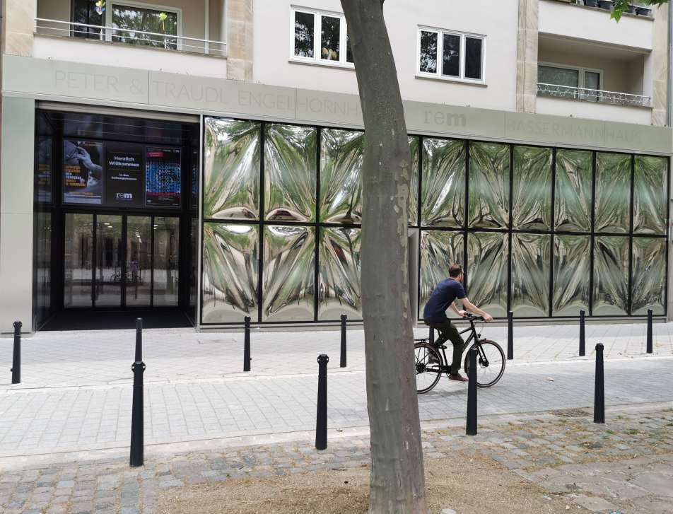 Fotografie in Farbe von einem Museum mit Glasfront und Eingangsbereich und einem Fahrradfahrer, der die Straße entlangfährt