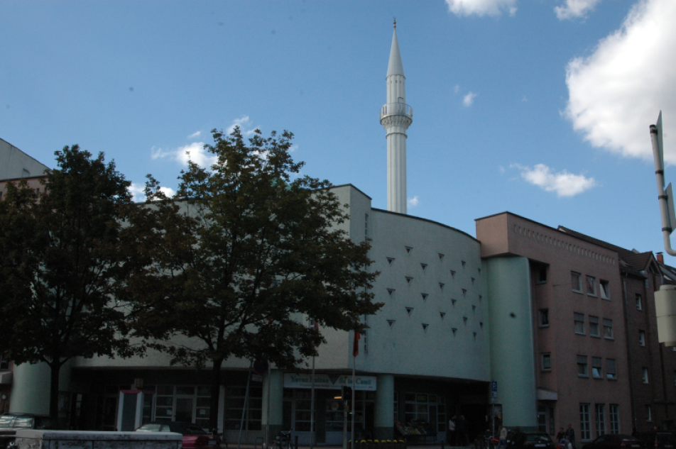 Farbfotografie von einer Moschee mit Bäumen im Vordergrund und leicht bewölktem Himmel