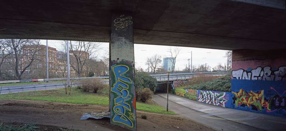Farbige Fotografie von einem Geh- und Fahrradweg unter einer Brücke mit Graffiti und Rasenflächen bei Tag